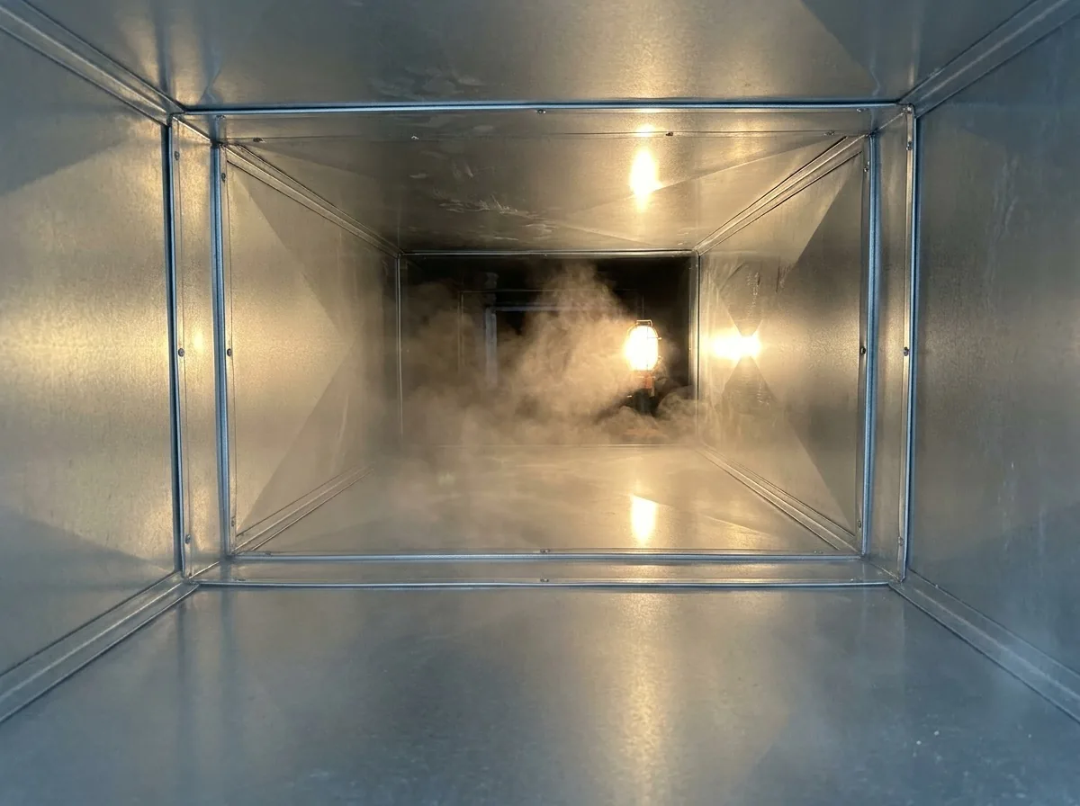 Looking along the clean sheet metal interior of a duct trunk line after cleaning, a fine sanitising mist drifting through and catching the work light