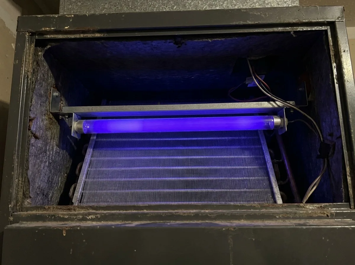 Germicidal UV lamp mounted above the evaporator coil inside an air handler, glowing deep violet blue with the coil fins lit below it